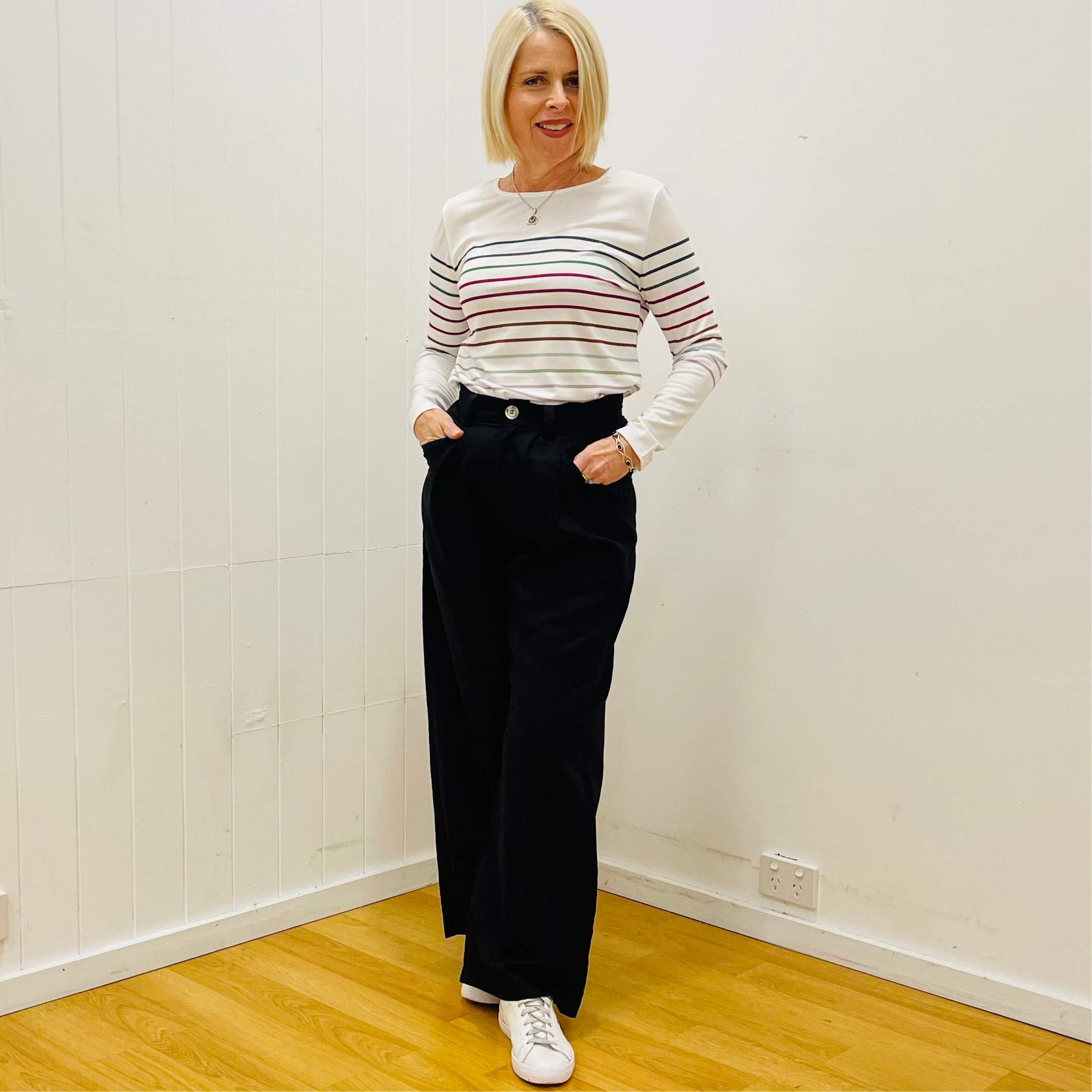 Woman wearing a striped shirt and black pants standing in a room with white walls and wooden floor.