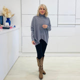 Grey knit sweater with long sleeves, high neckline and asymmetrical hemline. With black pants and brown embroidered boots.