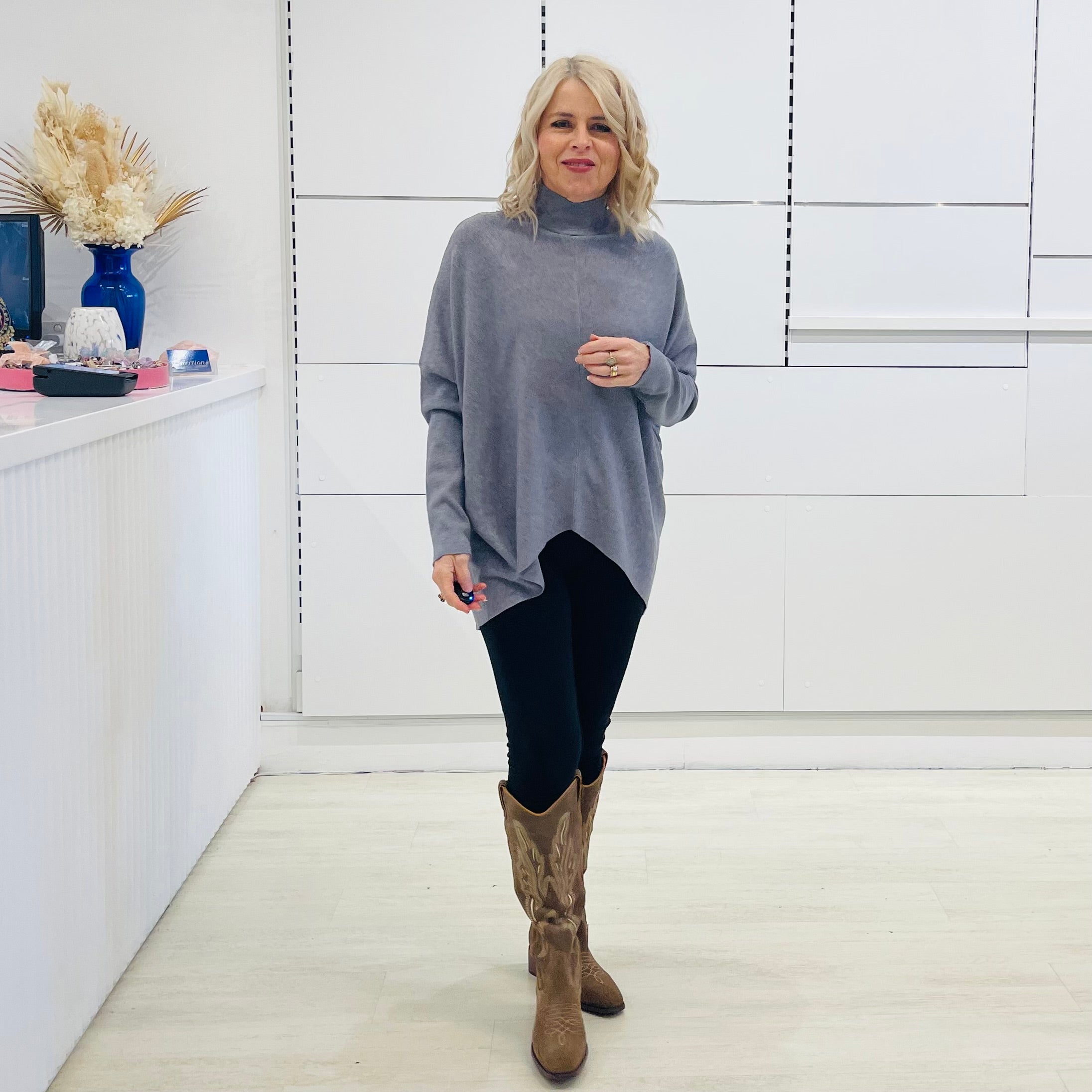Grey knit sweater with long sleeves, high neckline and asymmetrical hemline. With black pants and brown embroidered boots.