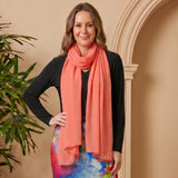 Woman wearing a colorful dress and pink scarf standing next to a potted plant against a beige wall.
