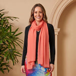 Woman wearing a colorful dress and coral scarf standing next to a potted plant against a beige wall.