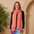 Woman wearing a colorful dress and coral scarf standing next to a potted plant against a beige wall.