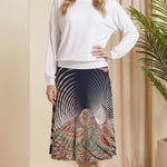 Woman wearing a white top and patterned skirt standing in a room with beige walls and a plant.