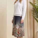 Woman wearing a white top and patterned skirt standing in a room with beige walls and a plant.