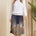 Woman wearing a white blouse and patterned skirt standing in a decorated room with beige walls and a plant.