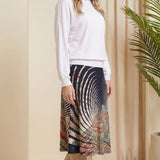 Woman wearing a white top and patterned skirt standing in a room with beige walls and a plant.