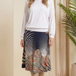Woman wearing a white top and patterned skirt standing in a room with beige walls and a plant.