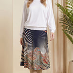 Woman wearing a white top and patterned skirt standing in a room with beige walls and a plant.
