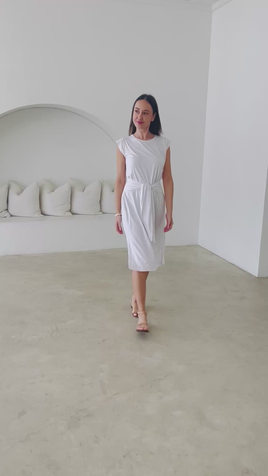 Woman in white dress with belt walking around simple room 