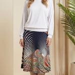 Woman wearing a white top and patterned skirt standing in a room with beige walls and a plant.