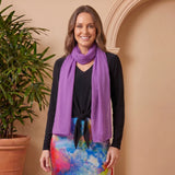 Woman wearing a colorful skirt and purple scarf standing next to a potted plant against a beige wall.
