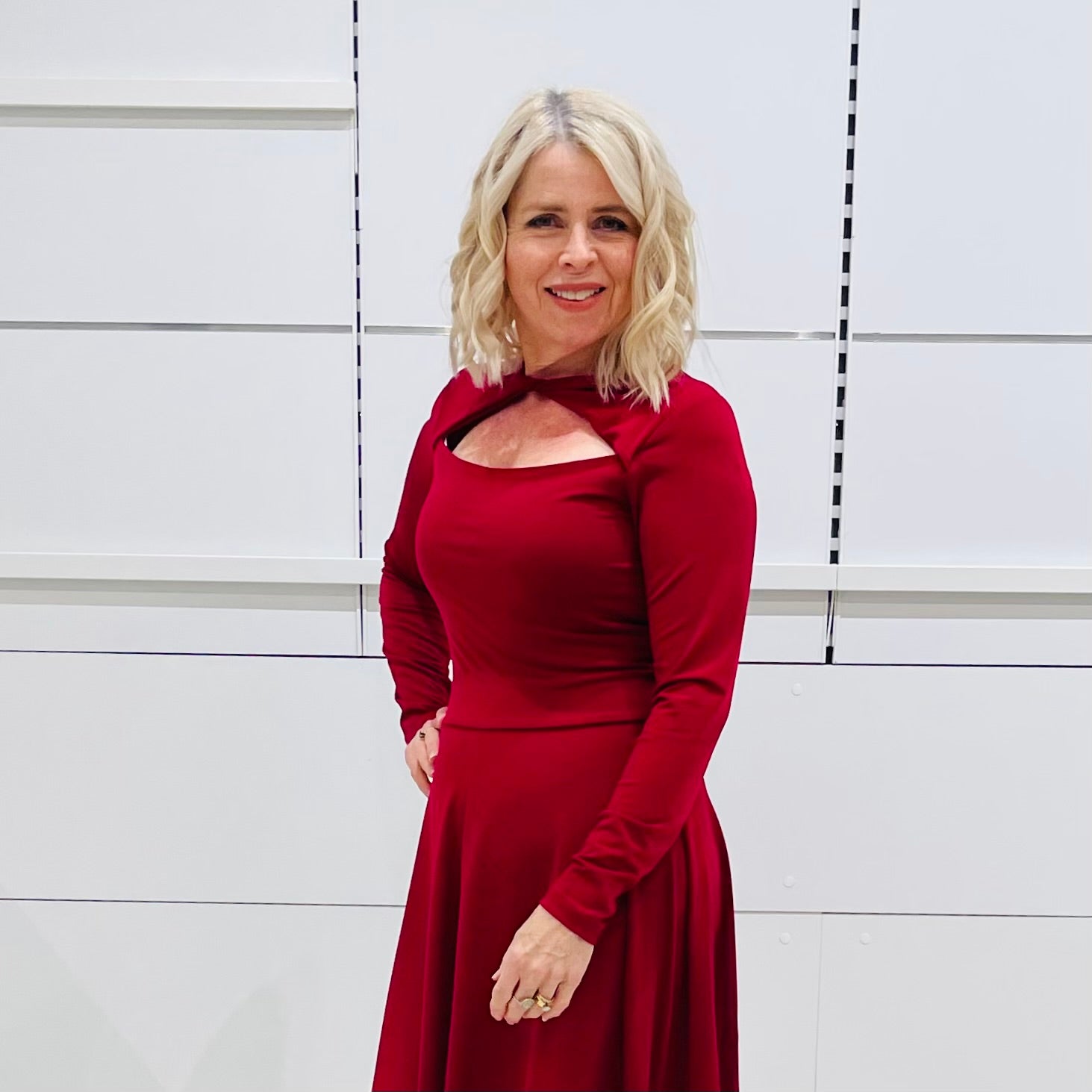 Close up, front view. Woman wearing the Penelope dress n rich ruby red, featuring a fit-and-flare, knee length, slihouette, full length sleeves, and a cut out neckline.